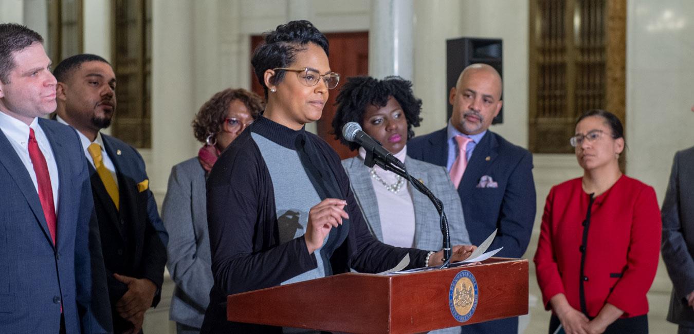 Representative Cephas speaking at podium in front of group of Democratic legislators