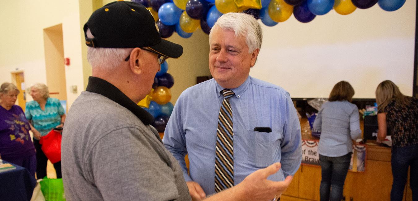 Representative Samuelson speaking to older man wearing veteran hat at indoor event