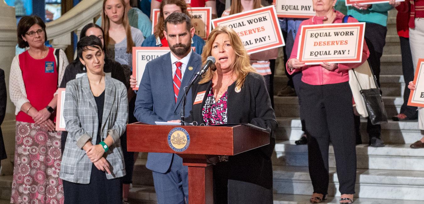 Representative Tina Davis speaking at podium in front of legislators and activists fighting for equal pay