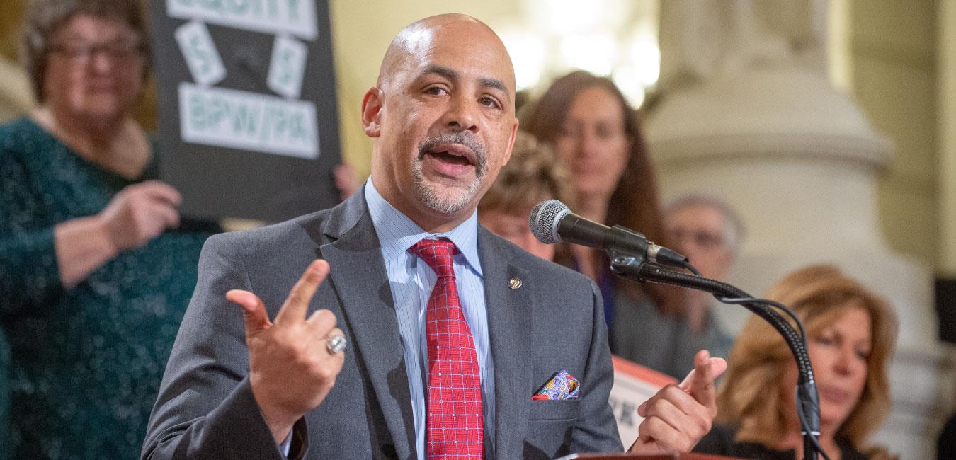 Representative Rabb holding up index fingers and speaking into microphone in front of group on Capitol rotunda steps