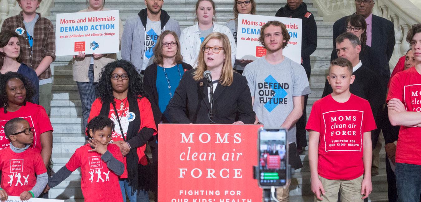 Representative Krueger speaking at podium with sign reading Moms Clean Air Force in front of activists on Capitol rotunda steps