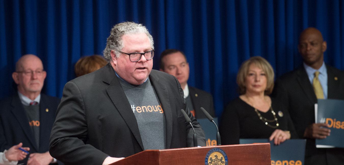 Representative Briggs speaking at podium in front of legislators wearing shirt reading #enough