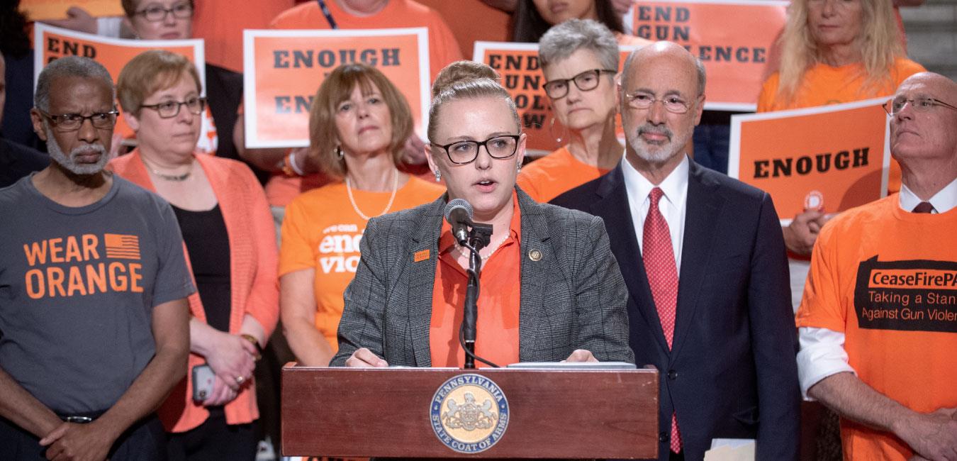 Representative O’Mara speaking at podium in front of Governor Wolf and gun safety advocates