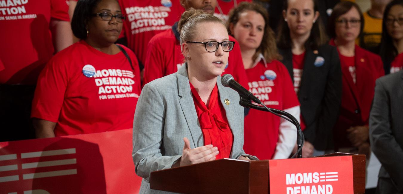 Representative O’Mara speaking at podium in front of Moms Demand Action members