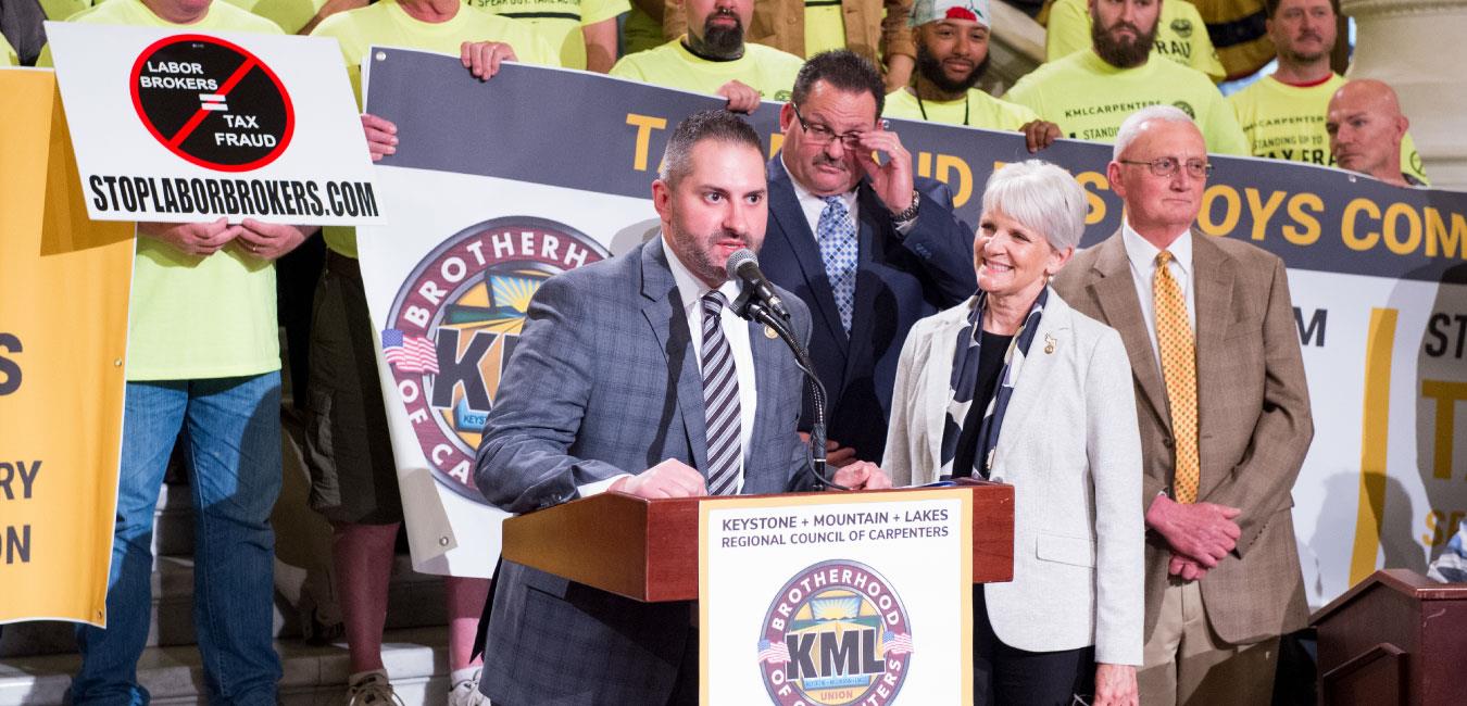 Representative Malagari standing at podium with lawmakers at Brotherhood of Carpenters rally in Capitol rotunda