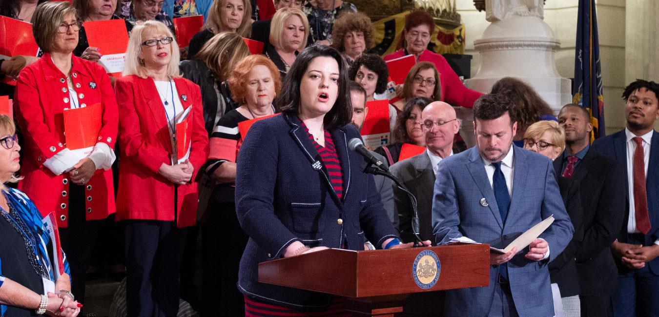 Representative Hanbidge speaking at podium next to fellow legislators and activists on Capitol rotunda steps